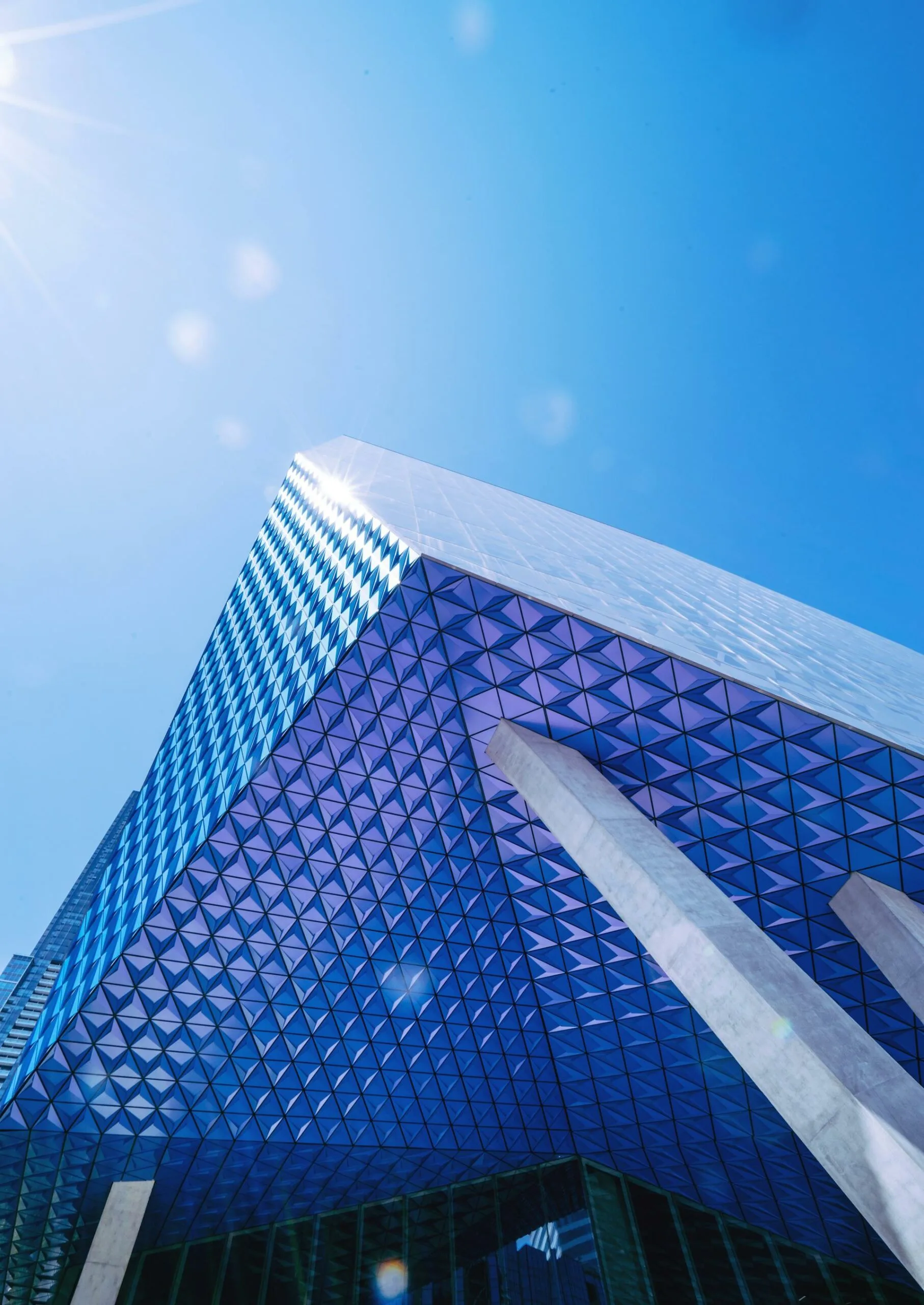 Looking up at a skyscraper with diamond-patterned triangular blue glass facade, concrete columns and a bright sun flare