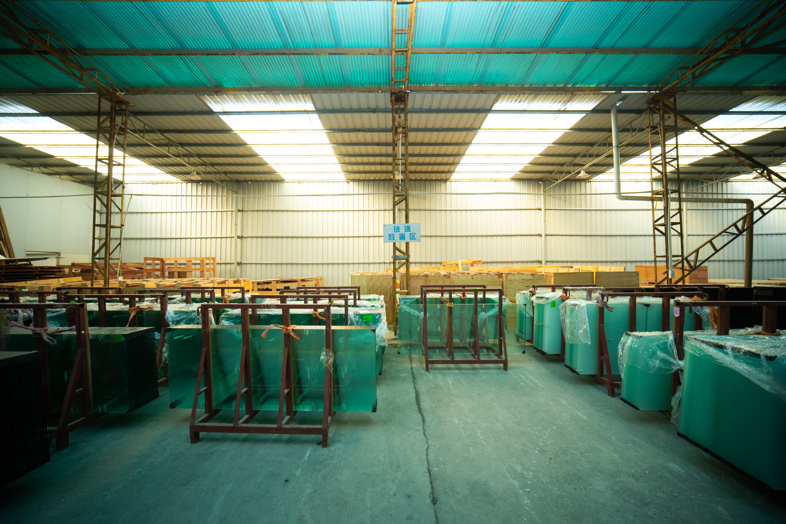Wrapped green tinted glass panels stored on red metal racks in the KJbath factory glass storage area