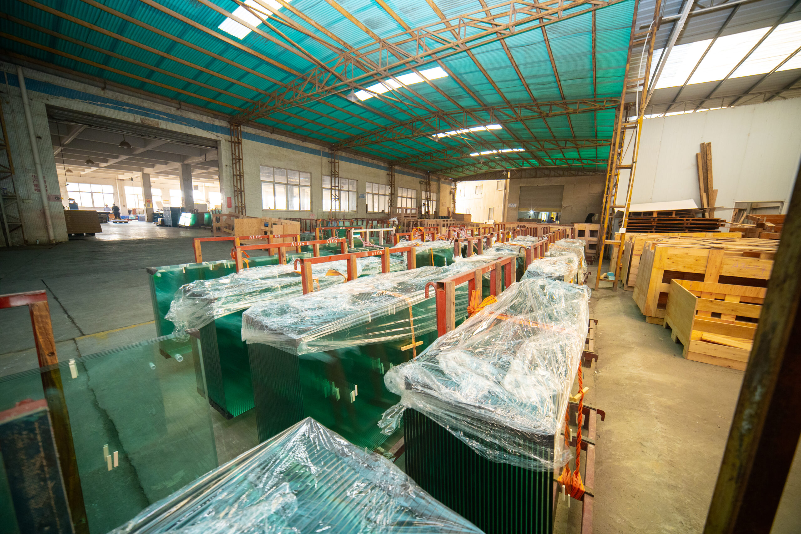 Rows of plastic-wrapped tempered glass panels on red racks in a factory glass warehouse