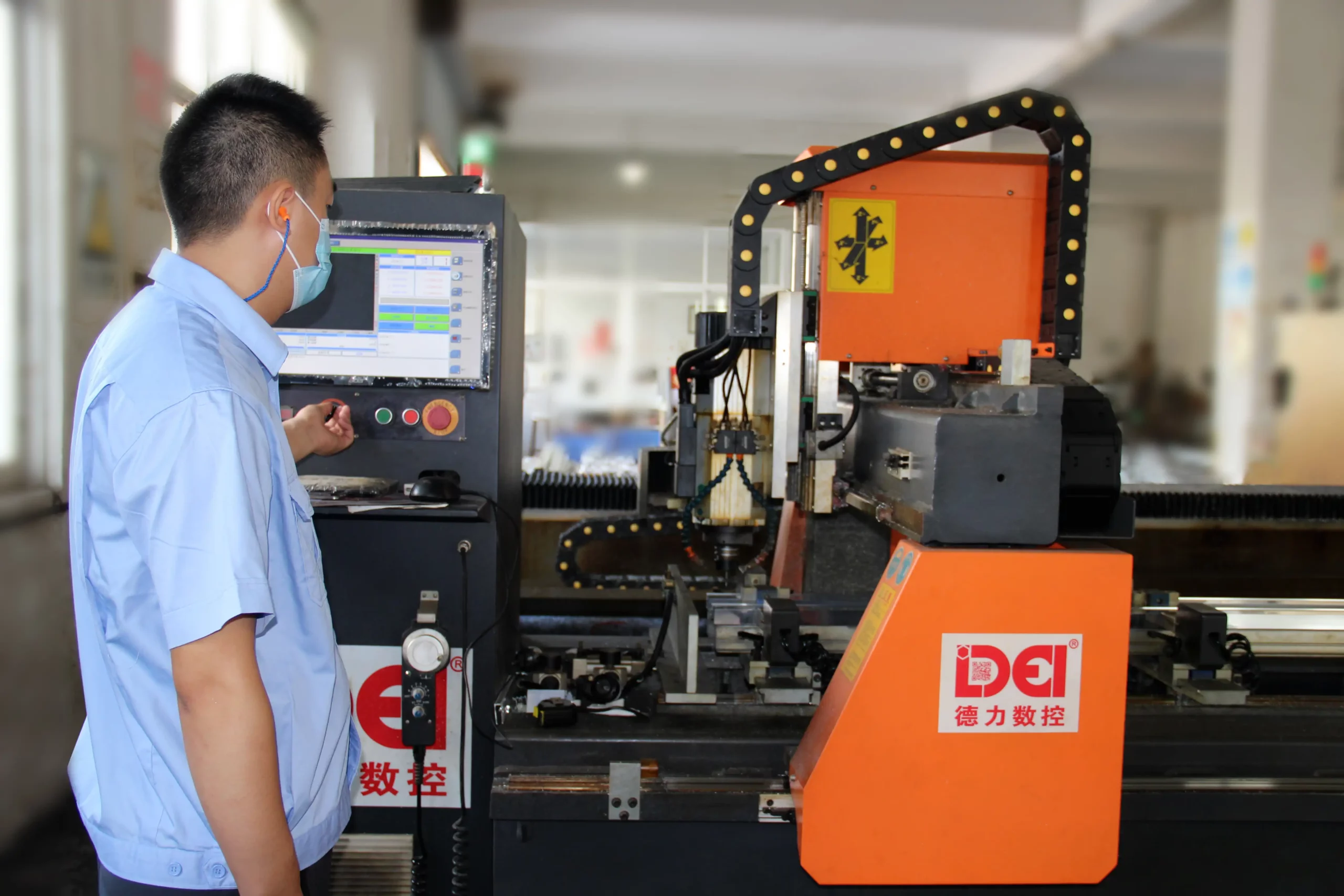 KJbath worker with mask and earplugs operating a DEI CNC machining center with orange housing and touchscreen panel