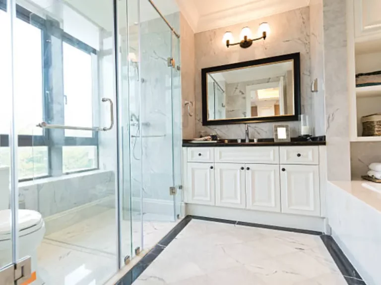 Frameless glass shower with chrome grab bar and bench in a marble bathroom with white vanity and black countertop
