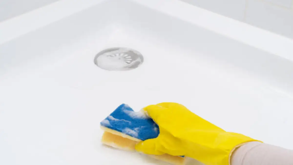 Hand in yellow rubber glove cleaning a white shower base surface with a soapy sponge