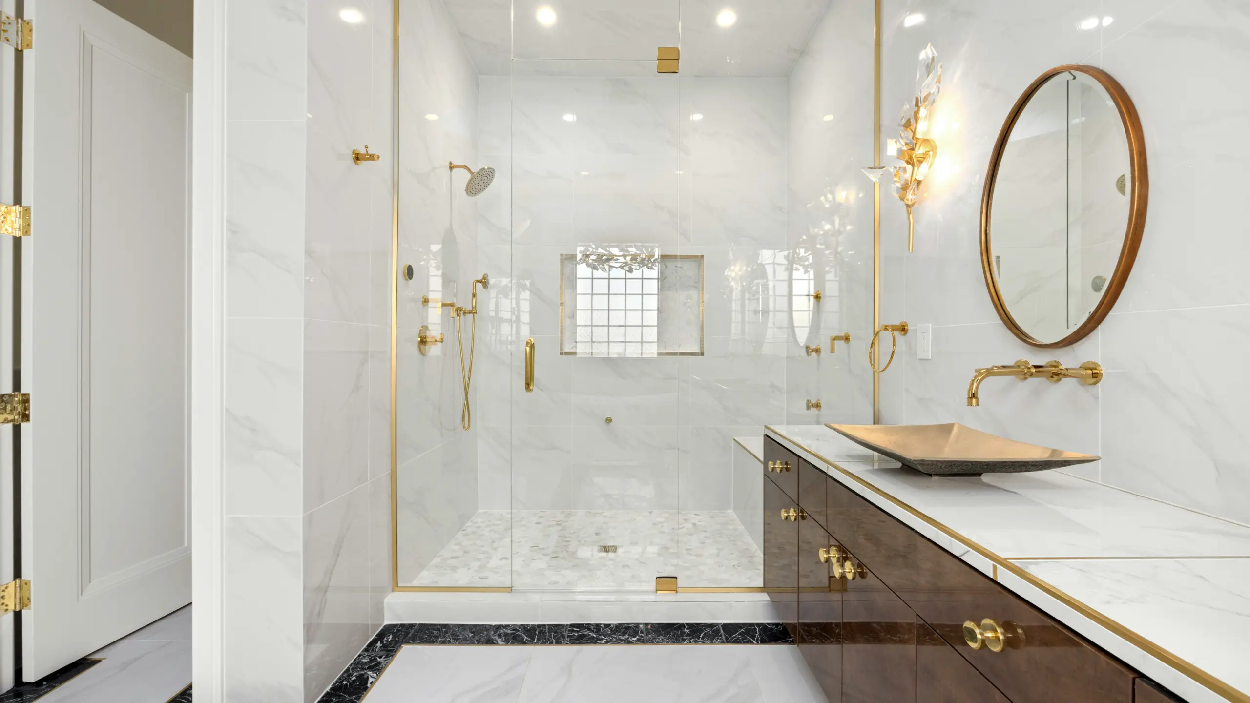 Elegant bathroom featuring white marble walls, gold brass fixtures, glass shower enclosure, dark wood vanity with vessel sink, and black marble floor accents