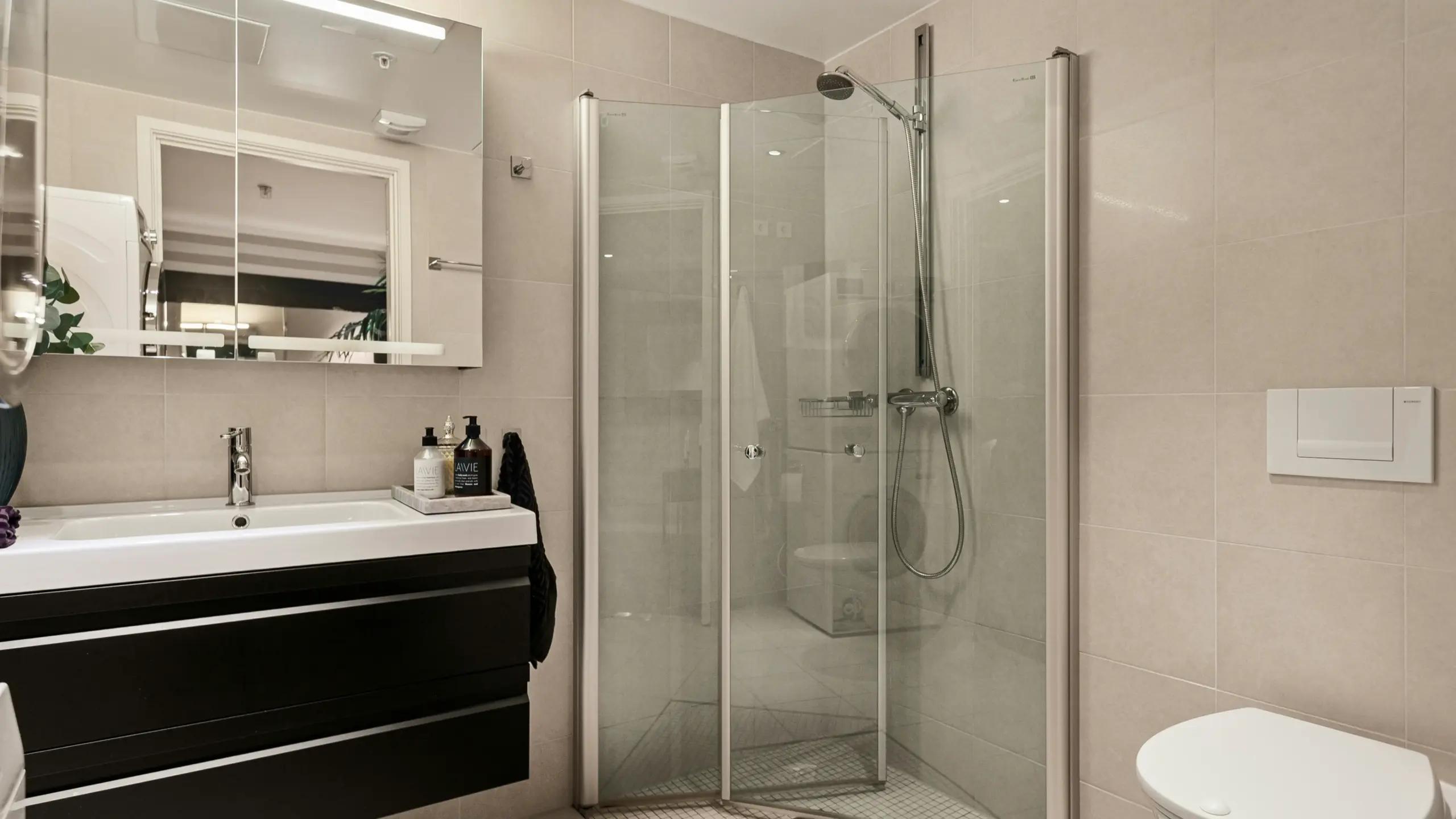 Contemporary bathroom featuring beige tiled walls, a frameless glass shower enclosure, floating black vanity with white sink, large mirrored cabinet, and wall-mounted toilet