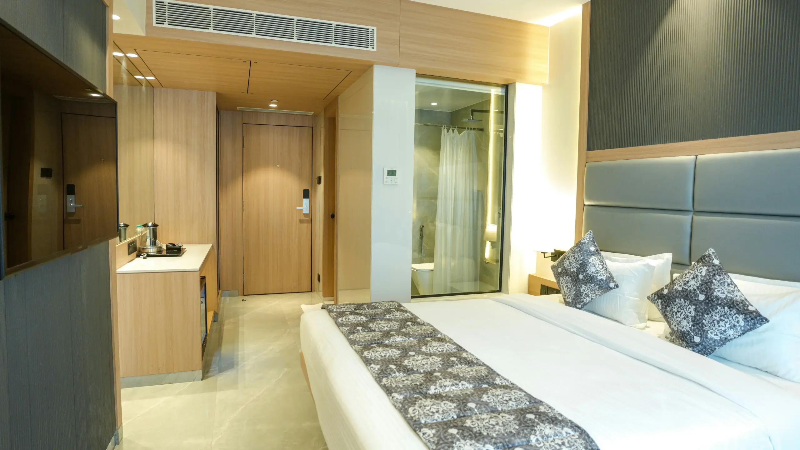 Interior of a modern hotel room featuring a double bed, glass-enclosed bathroom, and light wood finishes.