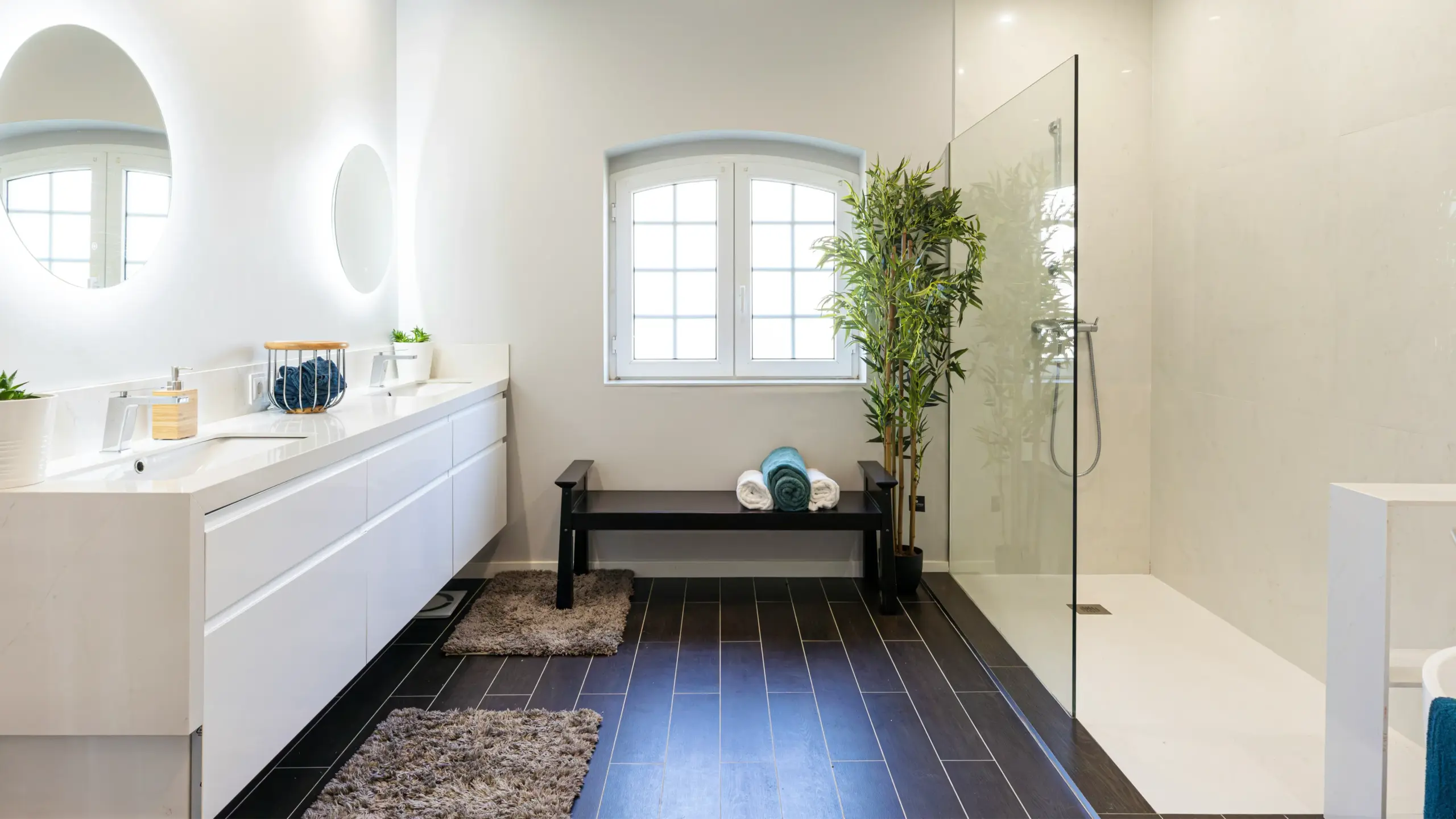 Bright modern bathroom with white double vanity, round mirrors, dark floor tiles, and glass shower enclosure.